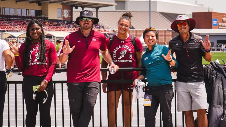 Five members of the NM State track and field team pose beside the track at the NCAA West First Round, with two wearing NM State track and field shirts and three wearing team apparel.