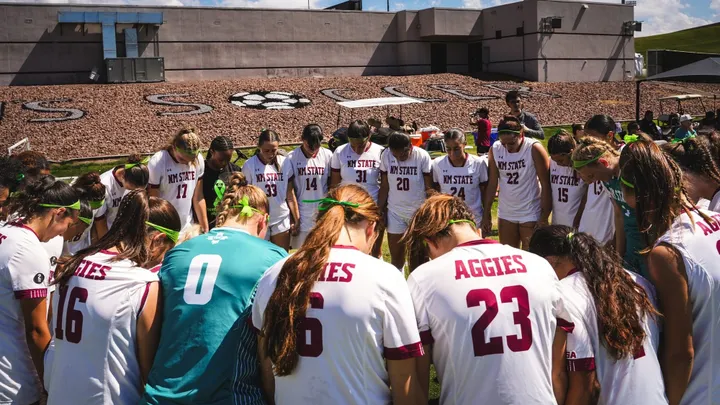 NM State women’s soccer players form a large circle on the field with their heads bowed before a match.