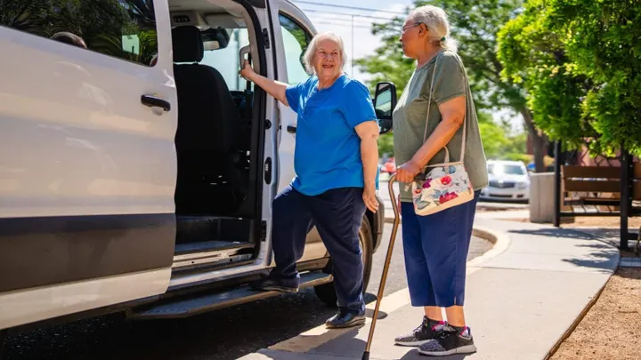 Two older women stand beside a van on a sidewalk.