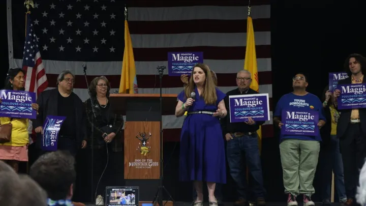 Maggie Toulouse Oliver speaks into a microphone at the New Mexico Democratic Party pre-primary convention as supporters hold campaign signs behind her.