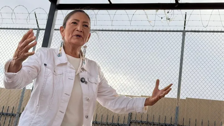 Democratic gubernatorial candidate Deb Haaland speaks with her arms extended outside Grand Summit Recovery and Workforce Development in Albuquerque, with a chain-link fence behind her.