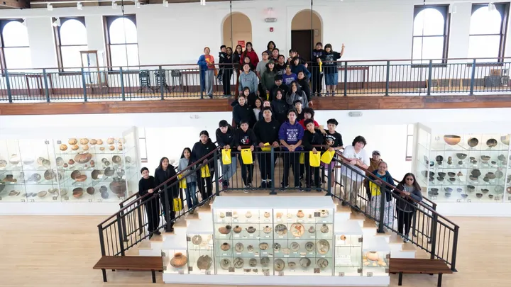 A large group of Mescalero Apache students and staff pose on a staircase and balcony inside the WNMU Museum, surrounded by display cases containing pottery and other artifacts.