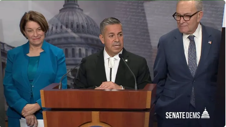 U.S. Sen. Ben Ray Luján speaks at a lectern during a Senate Democrats news conference, flanked by Sens. Amy Klobuchar and Chuck Schumer.