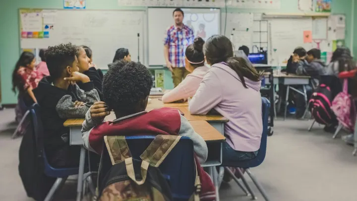 Students sit at classroom tables while a teacher speaks at the front of the room.