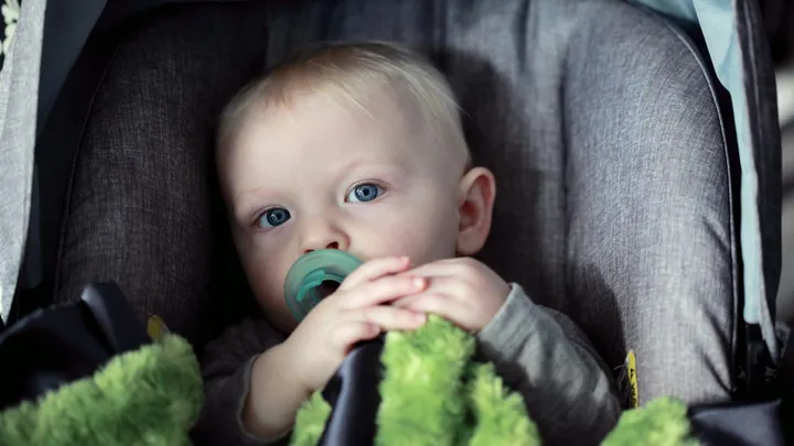 A baby with a pacifier sits in a gray child safety seat while holding a green blanket.