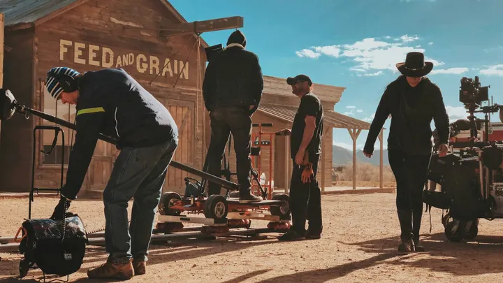 Film crew members set up camera equipment on a Western-style outdoor set.