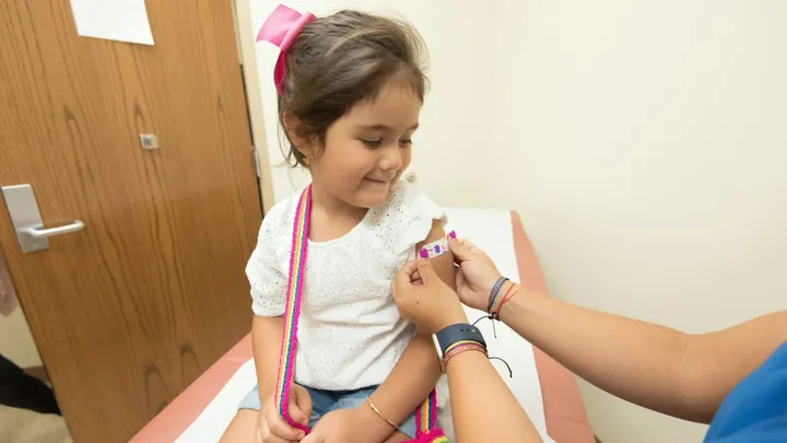 A child sits on an exam table as a health care worker places a bandage on her upper arm after a vaccination.