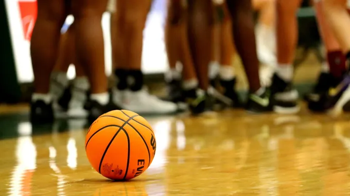 Basketball sits on a gym floor with players standing in the background.