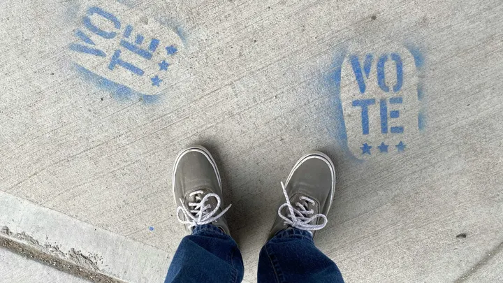 Two blue stenciled footprints reading “VOTE” are painted on a concrete walkway beside a voter’s shoes.