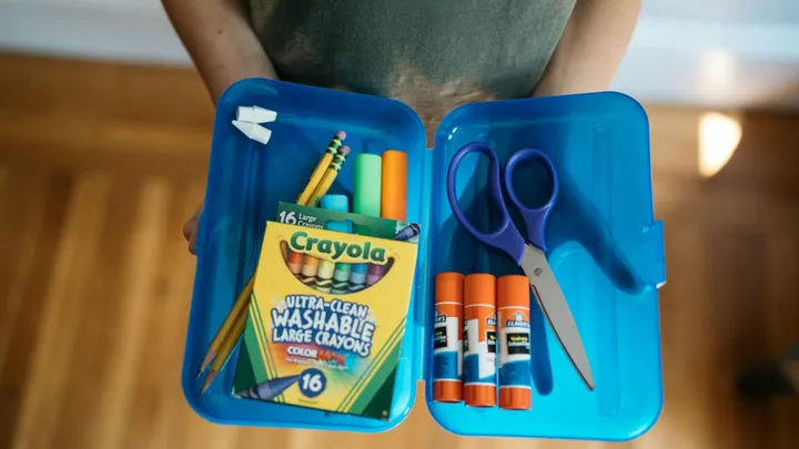 A person holds a blue pencil box filled with pencils, crayons, glue sticks and scissors.