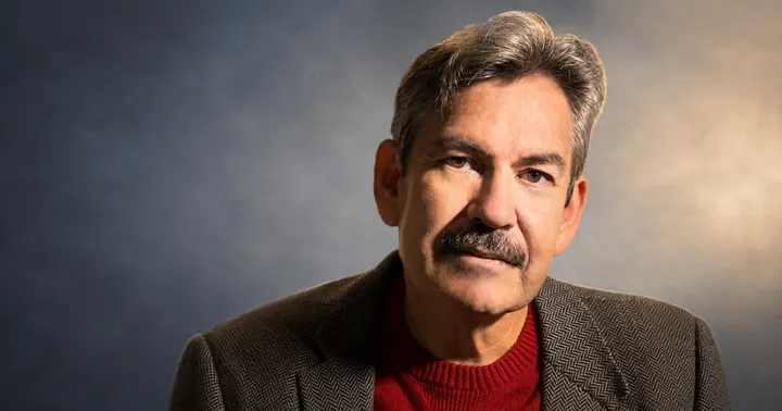 Benjamin Alire Sáenz, who died Tuesday at age 71, wears a dark herringbone jacket over a red sweater in a studio portrait.