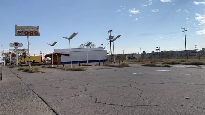 A vacant former gas station property with cracked pavement, overgrown weeds and old canopy structures is shown in Las Cruces.