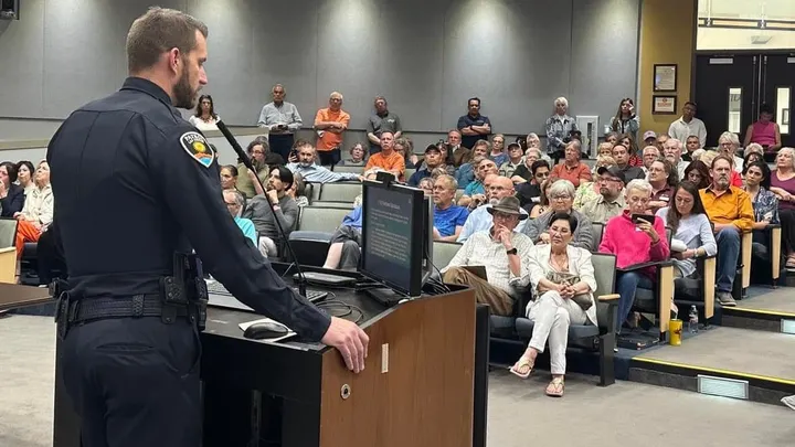 Las Cruces Police Chief Jeremy Story stands at a podium facing a crowded public meeting room.