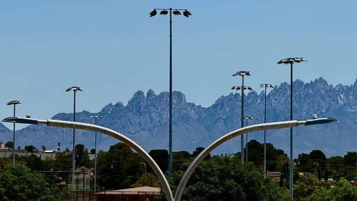Multiple light poles stand above a Las Cruces park with trees, buildings and the Organ Mountains in the background.