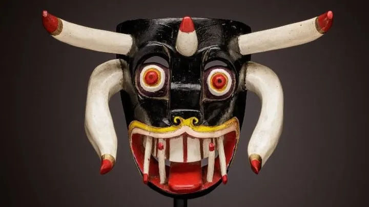 A black Mexican festival mask with horns, red and yellow eyes, white tusks and red details is displayed against a dark background.