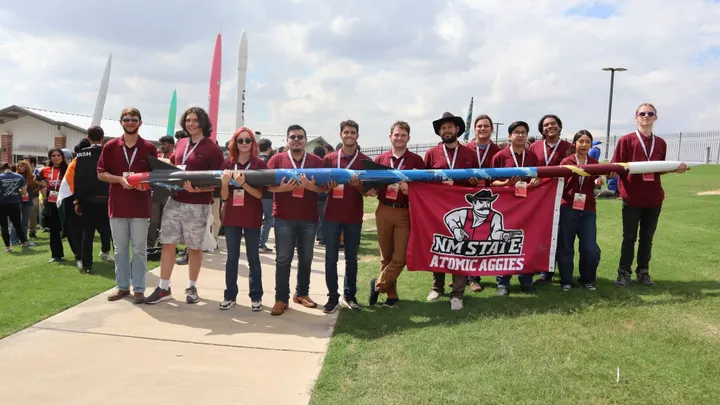 Members of NMSU’s Atomic Aggies stand beneath the 18-foot Crimson Comet II rocket while holding an NM State Atomic Aggies banner.