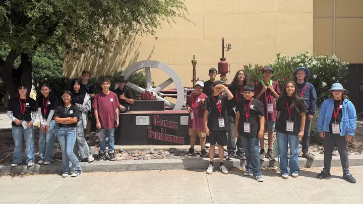 Middle school students pose for a group photo in front of a full-scale steam engine outside a building at NMSU.