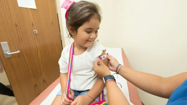 A young girl sits on an exam table as a health care worker places a colorful bandage on her upper arm after a vaccination.