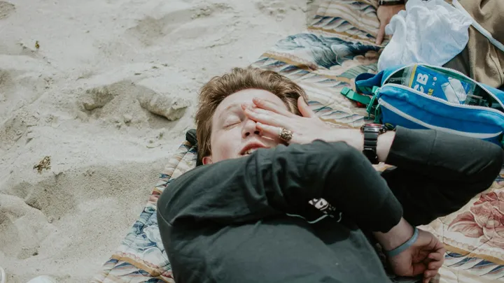 A person lies on a blanket outdoors with one hand covering their face during hot weather.