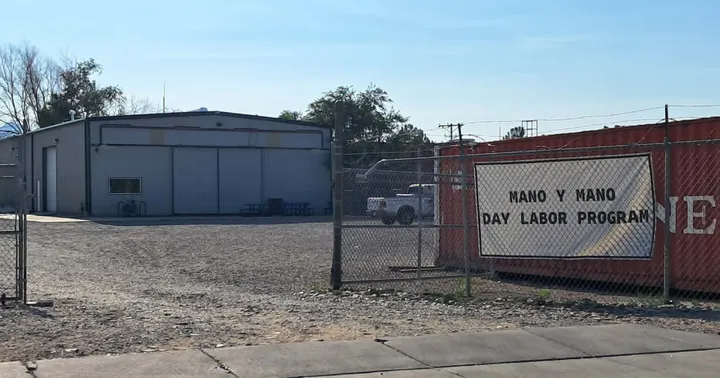 A fenced gravel lot and warehouse at Mesilla Valley Community of Hope in Las Cruces, with a sign for the Mano y Mano Day Labor Program.