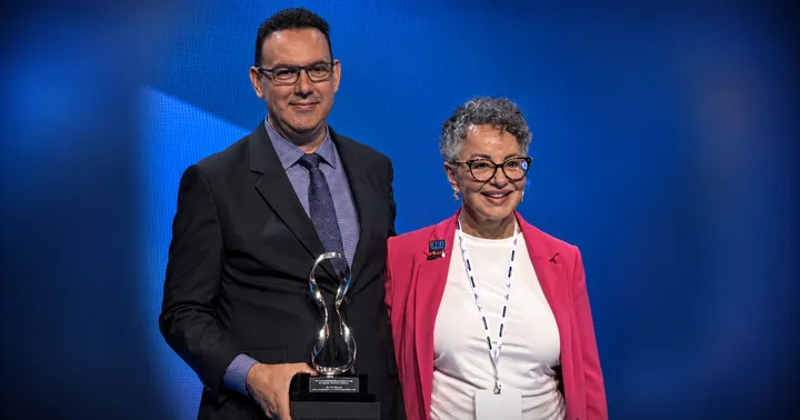 DACC professor Elmer González stands beside an event representative while holding the World Dental Hygienist Award against a blue stage backdrop.