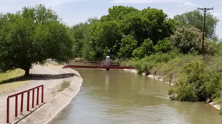 Water flows through a concrete-lined EBID irrigation canal bordered by trees and vegetation in southern New Mexico.