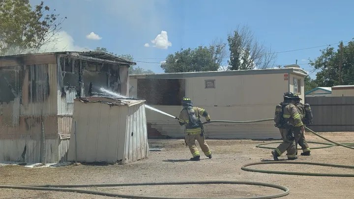Las Cruces firefighters spray water on a fire-damaged mobile home on Three Crosses Avenue while hoses lie across the ground.