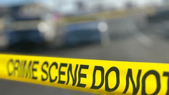 Yellow crime-scene tape stretches across the foreground, with police vehicles and a roadway blurred in the background.