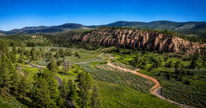 A sweeping Mimbres Valley landscape with green grasslands, pine-covered hills, red rock cliffs and a winding road beneath a clear blue sky.