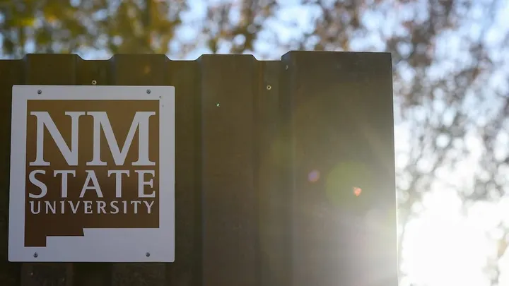 New Mexico State University logo mounted on a metal sign with sunlight shining through trees in the background.