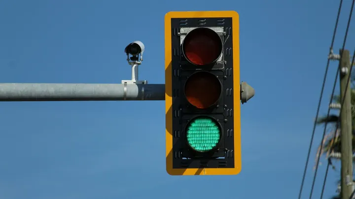 Traffic-enforcement camera mounted beside a green traffic signal against a clear blue sky.
