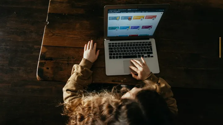A student uses a laptop at a wooden table with one hand on the trackpad and the other beside the computer.