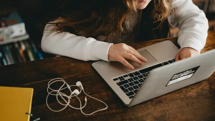 A student uses a laptop at a wooden table with earbuds and a notebook nearby.