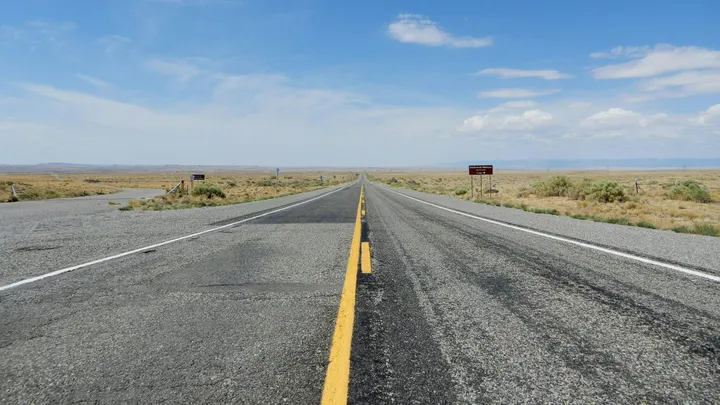 A two-lane highway stretches across a flat desert landscape beneath a blue sky in southern New Mexico.