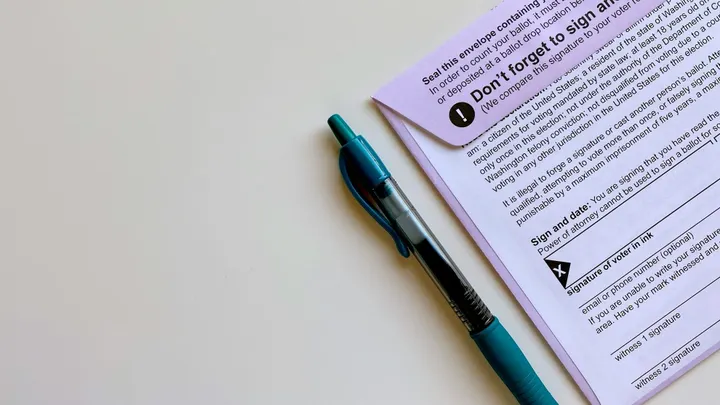 A mail ballot and blue pen rest on a table, with instructions and a signature line visible on the ballot envelope.