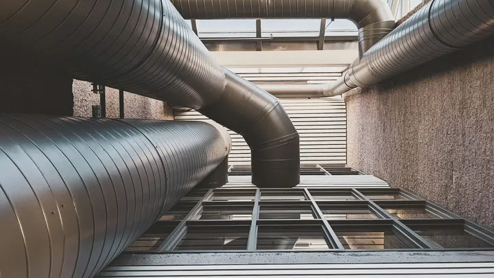 Large metal HVAC ducts run through an industrial-style building interior beneath a glass ceiling.