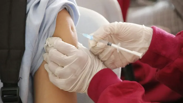 A health care worker wearing gloves administers a vaccine injection into a patient’s upper arm.