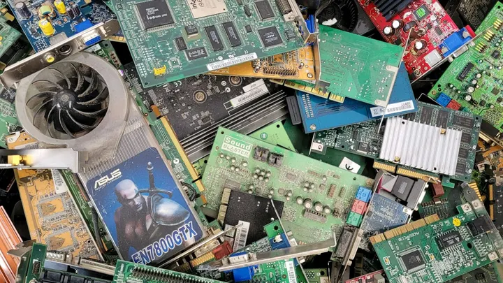 Pile of discarded computer circuit boards, graphics cards, fans and other electronic components stacked together.