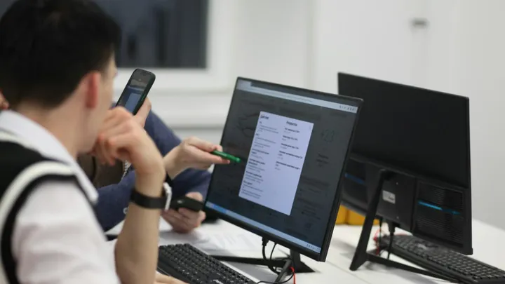 Students work at desktop computers and use smartphones in a classroom setting.