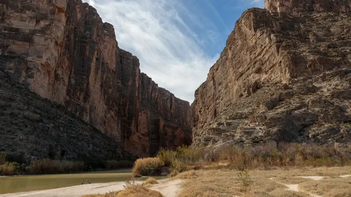 The Rio Grande runs through a rocky canyon beneath steep desert cliffs and a partly cloudy blue sky.