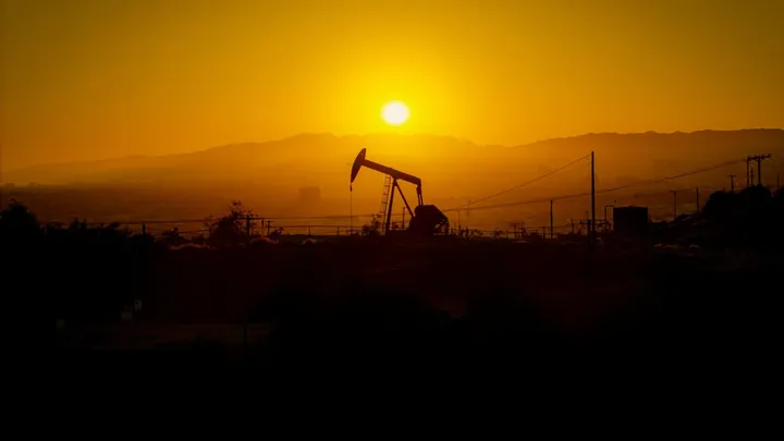 Silhouetted oil pumpjack and utility lines stand against an orange sunset over a desert landscape.