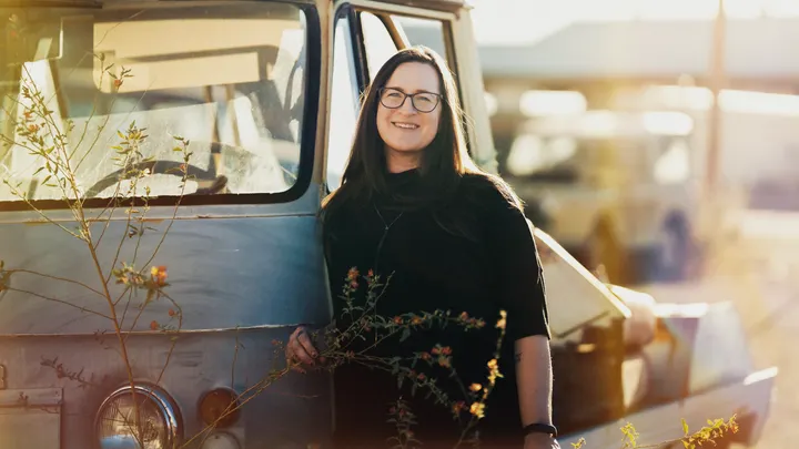 Athena Huckaby stands beside a weathered pickup truck outdoors, wearing glasses and a black top.