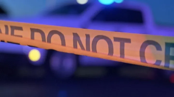 Orange police tape stretches across the foreground as blurred police vehicles flash blue lights in the background.