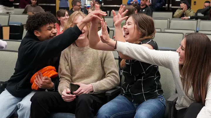 Four teens seated in an auditorium laugh and reach toward one another with raised hands during a group activity.