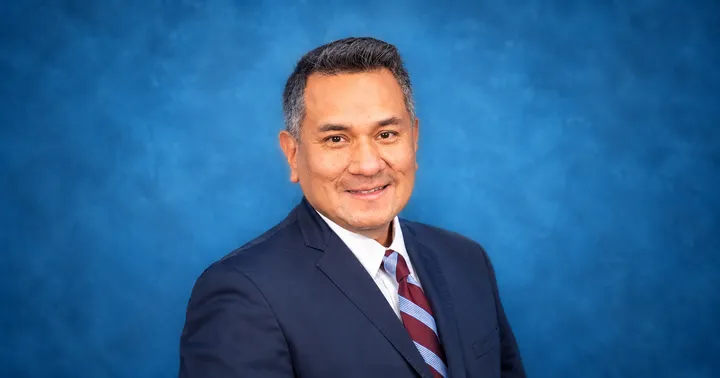 Las Cruces Mayor Pro Tem John P. Muñoz smiles in a suit and striped tie against a blue studio background.