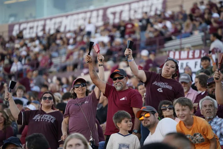 NM State football fans cheer from the stands at Aggie Memorial Stadium, many wearing crimson and holding cowbells.