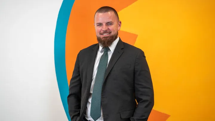 Logan Howard stands in a dark suit and green tie in front of a colorful orange, yellow and blue wall.