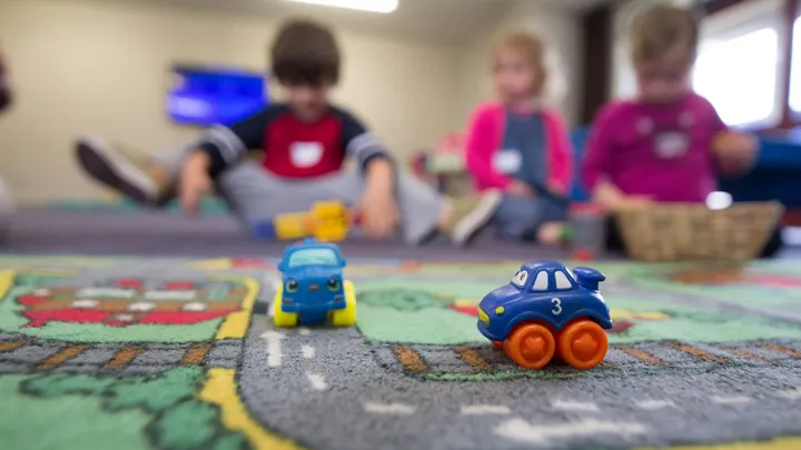 Toy cars sit on a colorful classroom rug while preschool-age children play in the background.