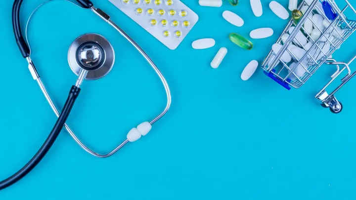 Stethoscope, assorted pills and a miniature shopping cart filled with medication sit on a bright blue background.