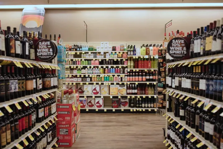 A grocery store alcohol aisle lined with bottles of wine and spirits on both sides, with shelves of canned drinks and boxed wine at the far end.